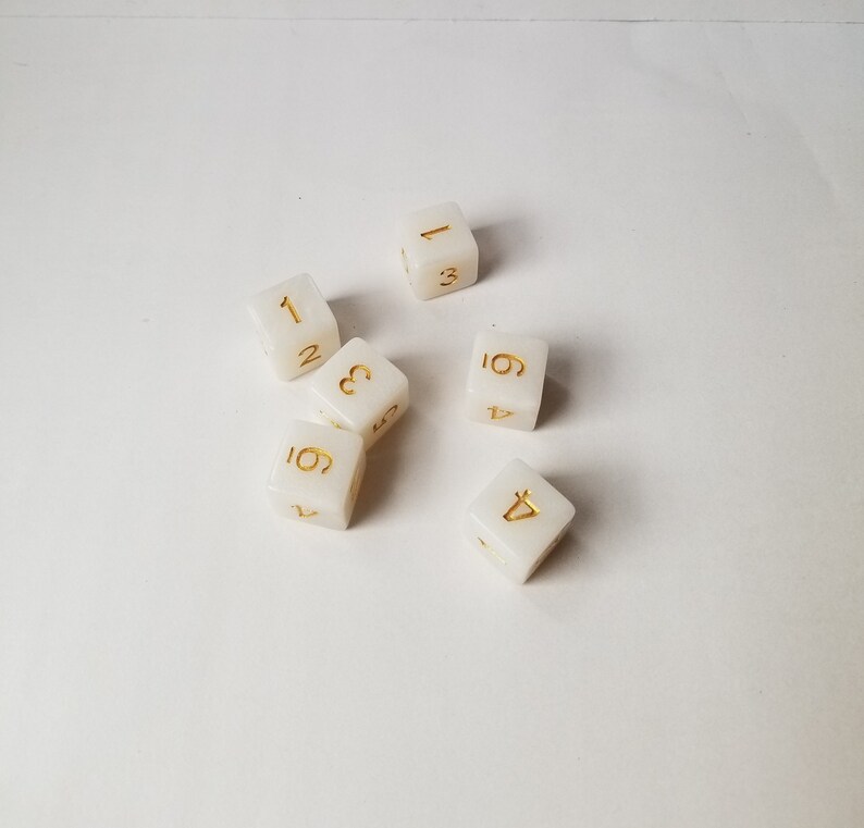 May include: Six white dice with gold numbers 1 through 6. The dice are scattered on a white surface.