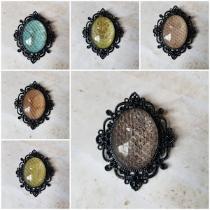 May include: Five black metal brooches with different colored glass insets. The brooches are oval shaped and have a decorative design around the glass. The glass insets are turquoise, yellow, brown, brown, and green.