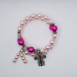 K&ouml;nnte beinhalten: Ein rosa-wei&szlig;es Perlenarmband mit einem silbernen Kreuzanh&auml;nger. Das Armband besteht aus kleinen rosa und wei&szlig;en Perlen und hat einen silbernen Kreuzanh&auml;nger.
