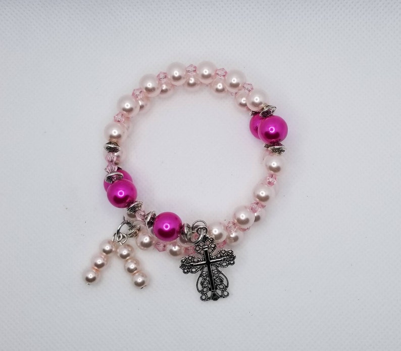 K&ouml;nnte beinhalten: Ein rosa-wei&szlig;es Perlenarmband mit einem silbernen Kreuzanh&auml;nger. Das Armband besteht aus kleinen rosa und wei&szlig;en Perlen und hat einen silbernen Kreuzanh&auml;nger.