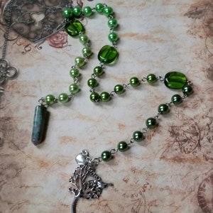 Op de afbeelding: Een groene kralenketting met een zilveren boomhanger en een groene edelsteenhanger. De ketting ligt op een getextureerde achtergrond.