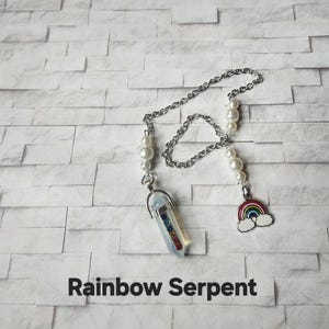 May include: A silver chain necklace with a rainbow crystal pendant and a rainbow charm. The necklace also features white pearl accents. The text "Rainbow Serpent" is printed in black at the bottom of the image.