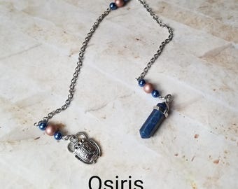 Puede incluir: Una cadena de plata con cuentas azules y de cobre, una punta de cristal azul y un dije de plata con forma de escarabajo egipcio. El texto "Osiris" está escrito debajo de la cadena.