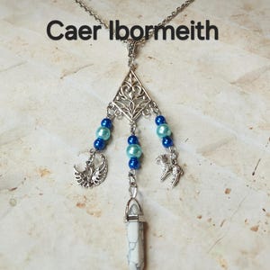 May include: Silver-toned pendant necklace with blue beads and charms. The necklace features a triangular filigree design, a white and gray stone pendant, and the text "Caer Ibormeith". Charms include a bird and a small animal.