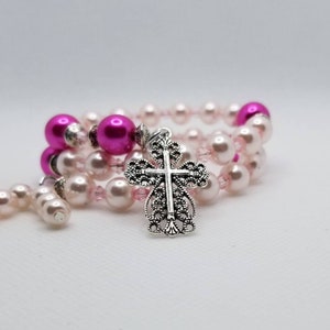 K&ouml;nnte beinhalten: Ein rosa und silbernes Perlenarmband mit einem silbernen Kreuzanh&auml;nger. Das Armband besteht aus kleinen rosa und wei&szlig;en Perlen und einer gr&ouml;&szlig;eren rosa Perle.