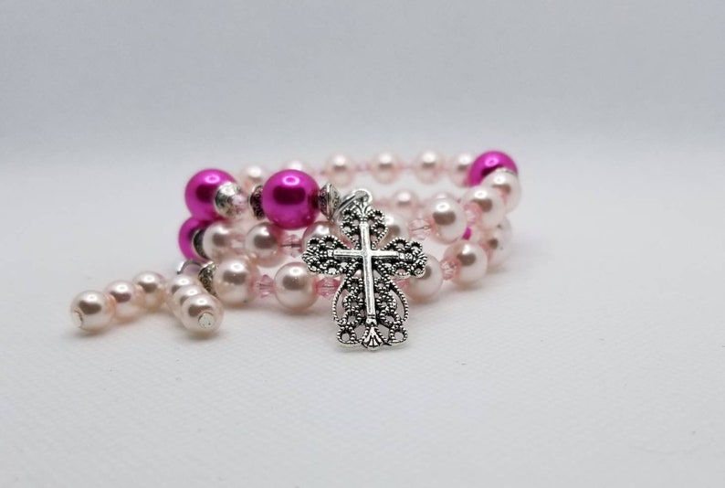 K&ouml;nnte beinhalten: Ein rosa und silbernes Perlenarmband mit einem silbernen Kreuzanh&auml;nger. Das Armband besteht aus kleinen rosa und wei&szlig;en Perlen und einer gr&ouml;&szlig;eren rosa Perle.
