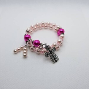 K&ouml;nnte beinhalten: Ein rosa-wei&szlig;es Perlenarmband mit einem silbernen Kreuzanh&auml;nger.