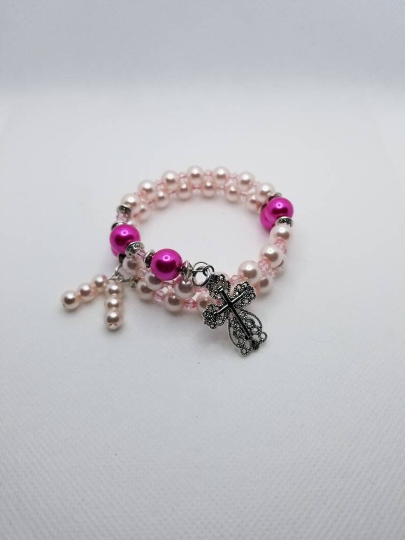 K&ouml;nnte beinhalten: Ein rosa-wei&szlig;es Perlenarmband mit einem silbernen Kreuzanh&auml;nger.