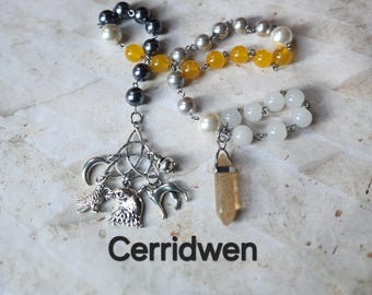 May include: A handmade necklace with a variety of beads in pearl, yellow, grey, and black. The necklace has a silver pendant with Celtic knot and animal charms, and a crystal pendant. The word "Cerridwen" is printed at the bottom.