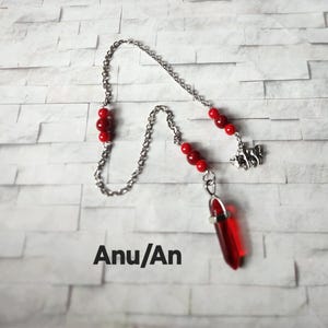 May include: A silver-toned metal chain pendulum featuring a faceted red crystal point, accented with small red beads and a silver-toned bull charm. The text "Anu/An" is printed in black near the bottom of the image, which displays a silver chain.