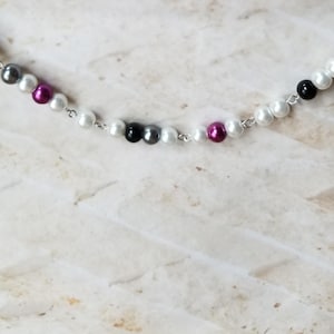 Peut inclure: Un collier délicat avec une chaîne argentée ornée de perles blanches, noires, grises et magenta alternées. Les perles sont espacées uniformément le long de la chaîne, créant un design simple mais élégant. Le collier est présenté sur une surface claire et texturée.