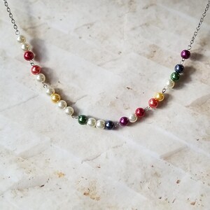 Op de afbeelding: Een zilveren ketting met een regenboog van gekleurde parels. De parels zijn wit, rood, geel, groen, blauw en paars.