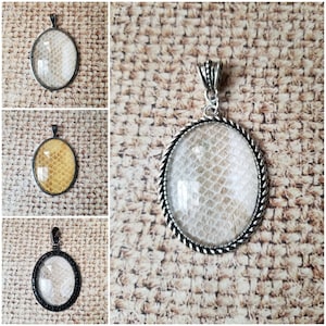 May include: Three silver pendant necklaces with oval-shaped glass inserts. The glass inserts are filled with a white, textured material that resembles snakeskin. The pendants are on a white, textured background.