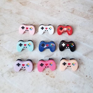 Op de afbeelding: Tien kleurrijke plastic gamecontroller-oorbellen. De oorbellen zijn in verschillende kleuren, waaronder roze, blauw, rood, wit en zwart. Elke oorbel heeft een hart in het midden van de controller.
