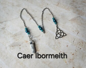 Puede incluir: Una cadena plateada con cuentas de color verde azulado y un colgante blanco y gris estampado. La cadena está conectada a un dije de nudo celta plateado. El texto "Caer Ibormeith" está impreso en negro en la parte inferior de la imagen.