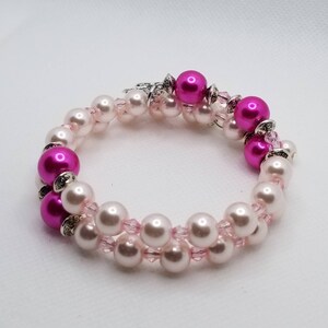 K&ouml;nnte beinhalten: Ein zweireihiges Armband mit rosa und wei&szlig;en Perlen. Das Armband hat einen silbernen Verschluss und ist mit kleinen rosa Kristallen verziert.