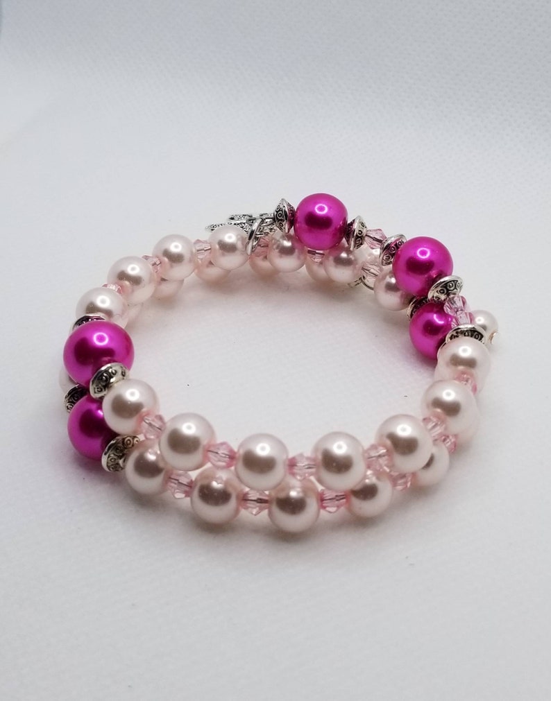 K&ouml;nnte beinhalten: Ein zweireihiges Armband mit rosa und wei&szlig;en Perlen. Das Armband hat einen silbernen Verschluss und ist mit kleinen rosa Kristallen verziert.