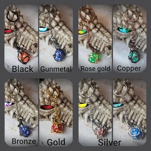 Puede incluir: Un conjunto de 10 llaveros de metal con un dado d20 enjaulado en varios colores, incluyendo negro, gris metalizado, oro rosa, cobre, bronce, oro y plata. Los llaveros se muestran en una estatua de dragón.