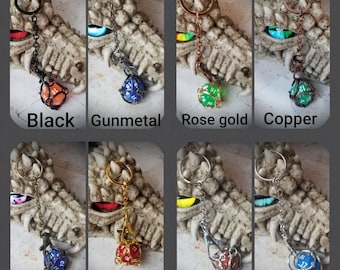 Op de afbeelding: Een set van 10 metalen sleutelhangers met een geketende d20 dobbelsteen in verschillende kleuren, waaronder zwart, gunmetal, rosé goud, koper, brons, goud en zilver. De sleutelhangers worden getoond op een drakenbeeld.