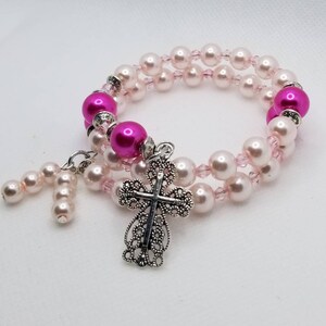 K&ouml;nnte beinhalten: Ein Armband mit zwei Reihen rosa und wei&szlig;en Perlen und einem silbernen Kreuzanh&auml;nger.