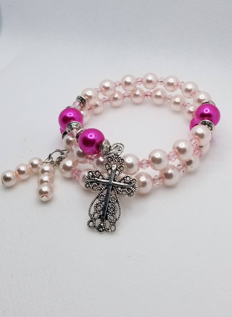 K&ouml;nnte beinhalten: Ein Armband mit zwei Reihen rosa und wei&szlig;en Perlen und einem silbernen Kreuzanh&auml;nger.