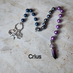 May include: A beaded necklace with dark blue, gray, and purple beads. The necklace has a silver charm with a star, a compass, and a crescent moon. A purple crystal pendant hangs at the bottom. The word "Crius" is printed below.