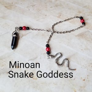 Op de afbeelding: Een zilveren ketting met zwarte en rode kralen, een zwarte kristallen hanger en een zilveren slangenbedel. De ketting is gelabeld met "Minoan Snake Goddess".