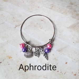 May include: A silver bangle bracelet with pink and purple beads and charms, including a seashell, a heart, and a starfish. The bracelet is labeled "Aphrodite".