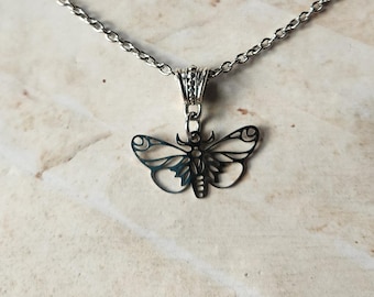 Op de afbeelding: Een zilverkleurige ketting met een delicate vlinderhanger. De hanger heeft gedetailleerde vleugelpatronen en hangt aan een fijne ketting. De ketting wordt gepresenteerd tegen een neutrale, getextureerde achtergrond, die het ontwerp van de sieraden benadrukt.