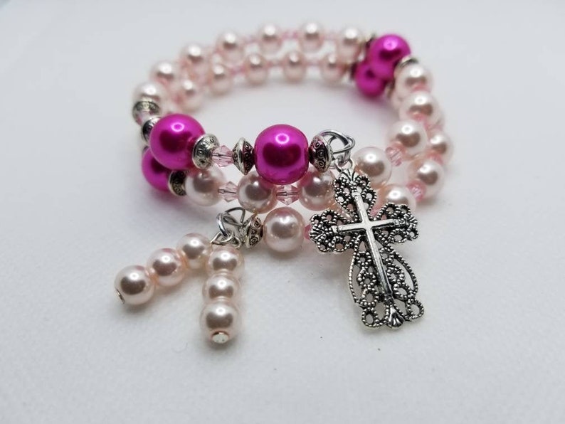 K&ouml;nnte beinhalten: Ein doppelreihiges Armband mit rosa und wei&szlig;en Perlen und einem silbernen filigranen Kreuzanh&auml;nger. Das Armband hat einen Anh&auml;nger mit rosa Perlen.