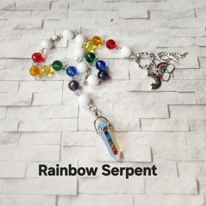 May include: A necklace with white and rainbow-colored beads, and a crystal pendant. The pendant is a clear, elongated shape with rainbow-colored stripes. The necklace also features charms of a rainbow, moon, and clouds. The text "Rainbow Serpent" is at the bottom.