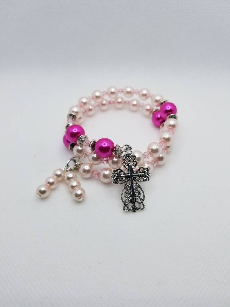 K&ouml;nnte beinhalten: Ein rosa-wei&szlig;es Perlenarmband mit einem silbernen Kreuzanh&auml;nger. Das Armband besteht aus kleinen, runden Perlen und hat einen zarten, femininen Look.