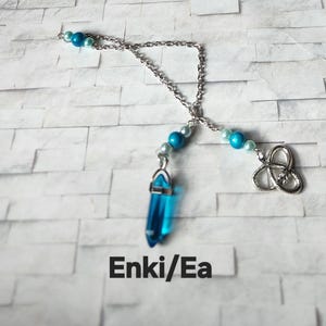 Könnte beinhalten: Eine silberfarbene Kettenhalskette mit hellblauen und dunkelblauen Perlen, einem durchscheinenden blauen Kristallanhänger und einem silberfarbenen Triquetra-Anhänger. Der Text Enki/Ea steht unter dem Schmuckstück auf einer weißen Ziegeloberfläche.