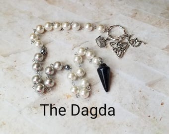 May include: A white pearl and silver pendulum with a black crystal point, a crown charm, a Celtic knot charm, and a crescent moon charm. The text "The Dagda" is written below the pendulum.