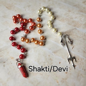 May include: A beaded necklace with red, orange, and cream-colored beads. A red and white pendant hangs from the red beads. The necklace has a silver-colored trident charm. The text "Shakti/Devi" is below the necklace.