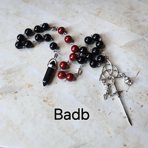 Peut inclure: Un chapelet avec des perles noires et rouges, un pendentif en cristal noir et un charm argenté avec une épée et un nœud celtique. Le mot "Badb" est imprimé en noir en bas.