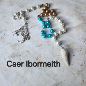 May include: A collection of beaded necklaces in various colors, including white, gold, and turquoise. One necklace features a silver pendant with Celtic knot and crescent moon designs. The text "Caer Ibormeith" is displayed at the bottom.