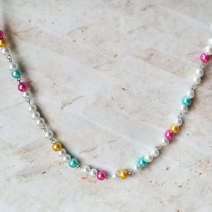 Peut inclure: Un collier délicat avec une chaîne argentée et des perles blanches, roses, jaunes et turquoise. Les perles rondes créent un accessoire coloré et élégant. Un bijou parfait pour toutes les occasions.