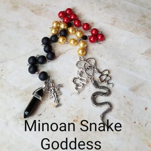 Op de afbeelding: Een zwarte, gouden en rode kralenketting met een zwarte kristallen hanger en een zilveren slangenbedel. De ketting is gelabeld "Minoïsche Slangengodin".