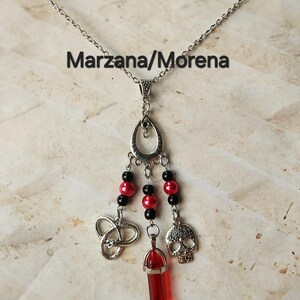May include: Silver-toned pendant necklace with a chain. The pendant features a teardrop-shaped top with dangling elements: black and red beads, a Celtic knot, a red crystal, and a skull charm. The text "Marzana/Morena" is displayed.