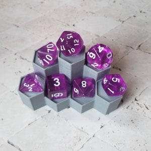 May include: A gray plastic hexagonal dice tray with seven compartments holds eight purple dice with white numbers. The dice are numbered 1-6, 8, 10, 12, 15, 17, 23, 32, 59, 60, 70.