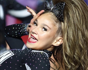 May include: A young cheerleader in a black uniform with silver accents and sparkling embellishments. She wears a black bow with rhinestones in her hair and has red lipstick. The image captures a moment of performance.