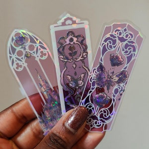 May include: Three iridescent bookmarks with a purple and white color scheme. The bookmarks feature intricate designs of flowers, butterflies, and a castle.