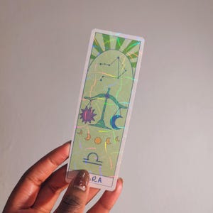 May include: A rectangular Libra-themed bookmark with a light green background and holographic accents. The design includes a constellation, scales, a sun, and a crescent moon. The word "RA" is at the bottom.