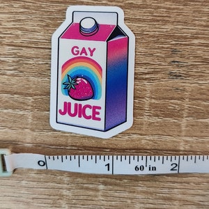 Puede incluir: Una pegatina de una caja de zumo gay con un arco&iacute;ris y una fresa en la parte delantera. La caja es rosa, azul y morada.