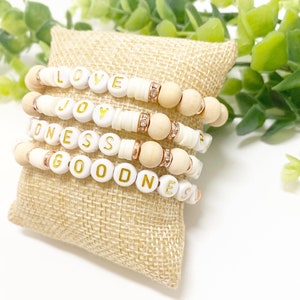 May include: Four stacked bracelets with white and gold letter beads spelling "LOVE", "JOY", "KINDNESS", and "GOODNESS". The bracelets also feature wooden beads and rose gold accents, displayed on a burlap pillow.