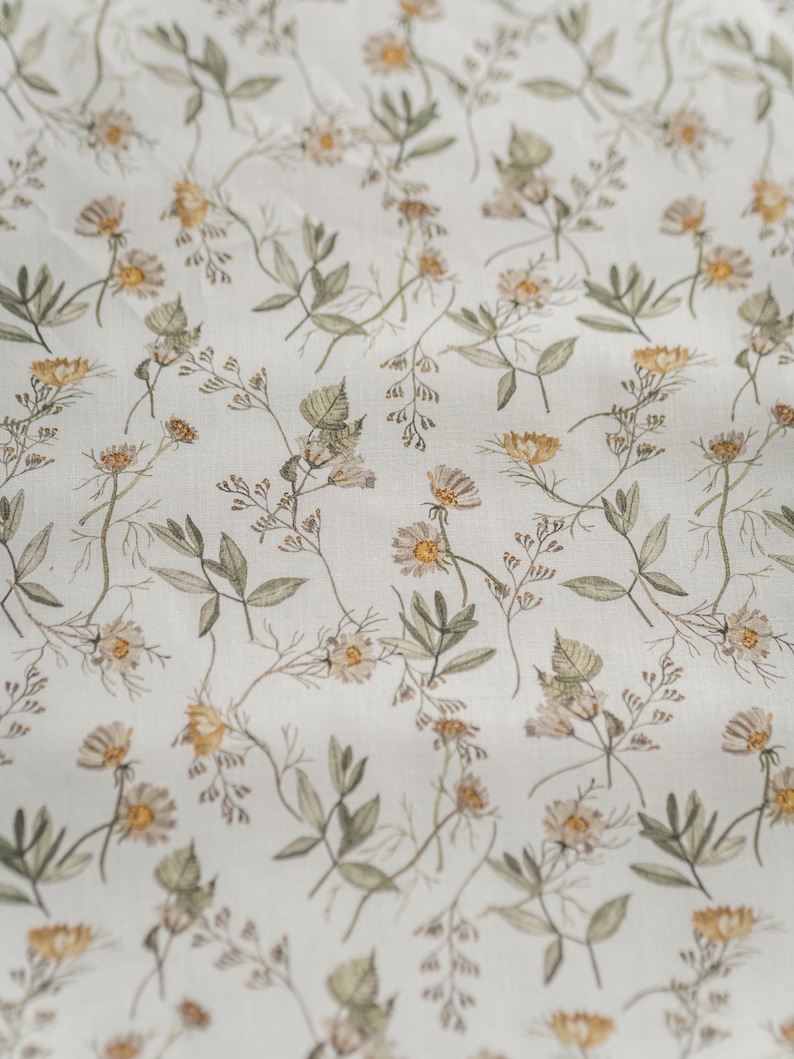 May include: A white fabric with a delicate floral pattern featuring small, light brown and yellow flowers and green leaves.