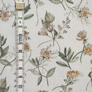 May include: White fabric with a repeating pattern of small, light brown and beige flowers and leaves. A white measuring tape with black markings is on the left side of the image.