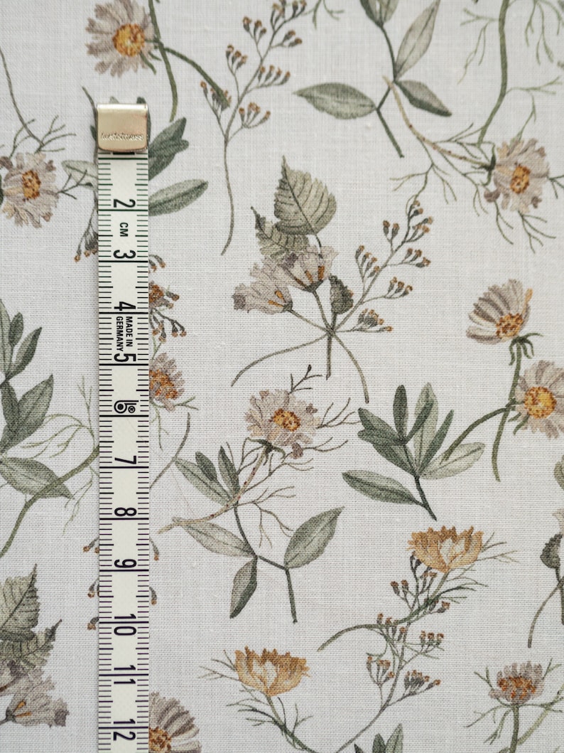 May include: White fabric with a repeating pattern of small, light brown and beige flowers and leaves. A white measuring tape with black markings is on the left side of the image.