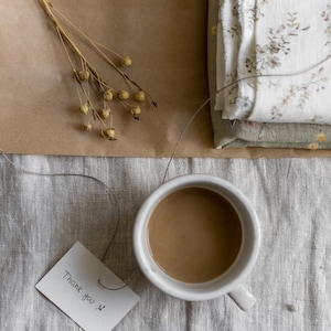Puede incluir: Una taza de cer&aacute;mica blanca llena de caf&eacute; se encuentra sobre un mantel de lino beige. Una peque&ntilde;a tarjeta blanca con el texto "Gracias" est&aacute; sobre la mesa junto a la taza.